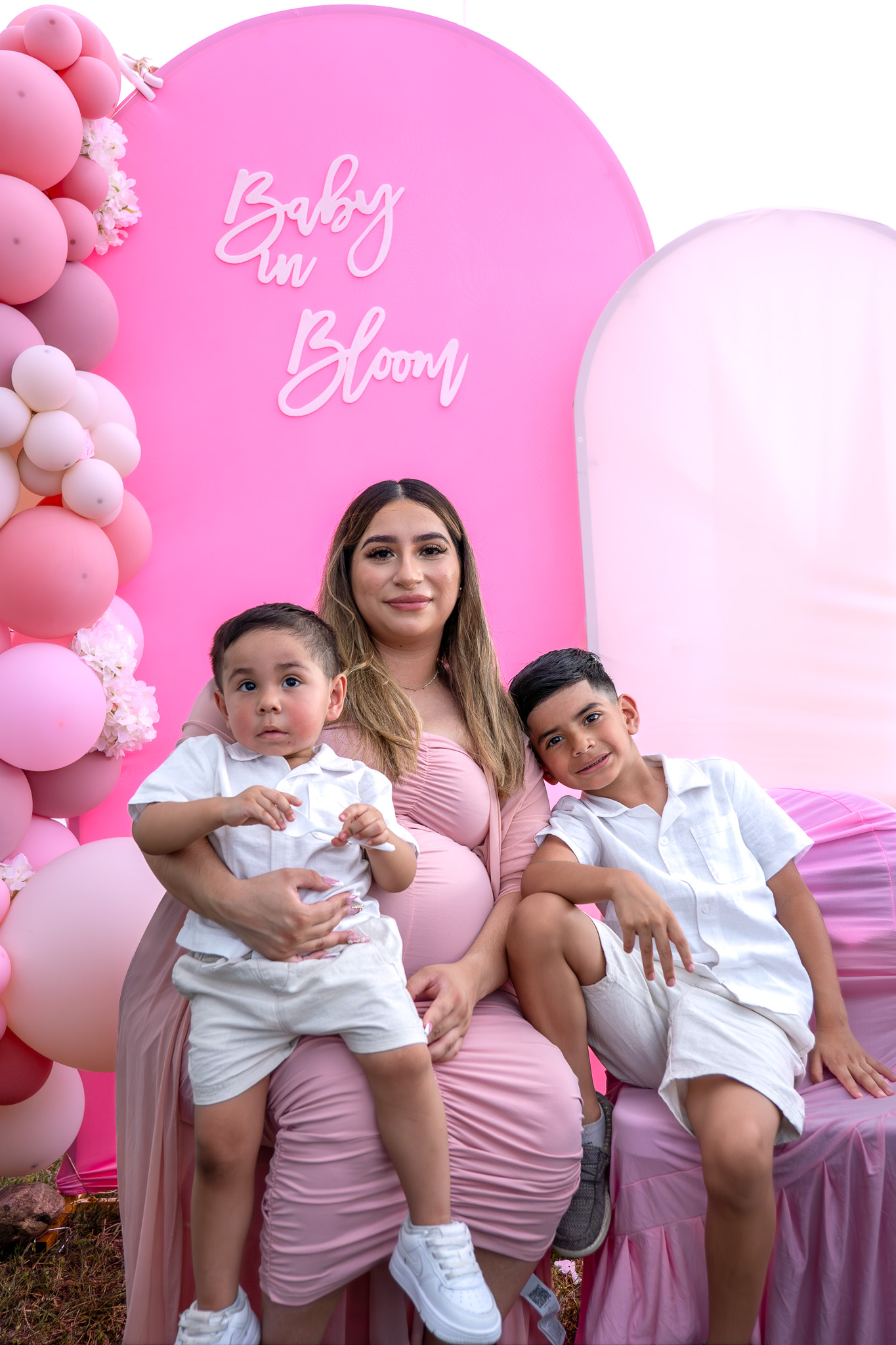 Expecting mother with her two sons at a pink baby shower backdrop